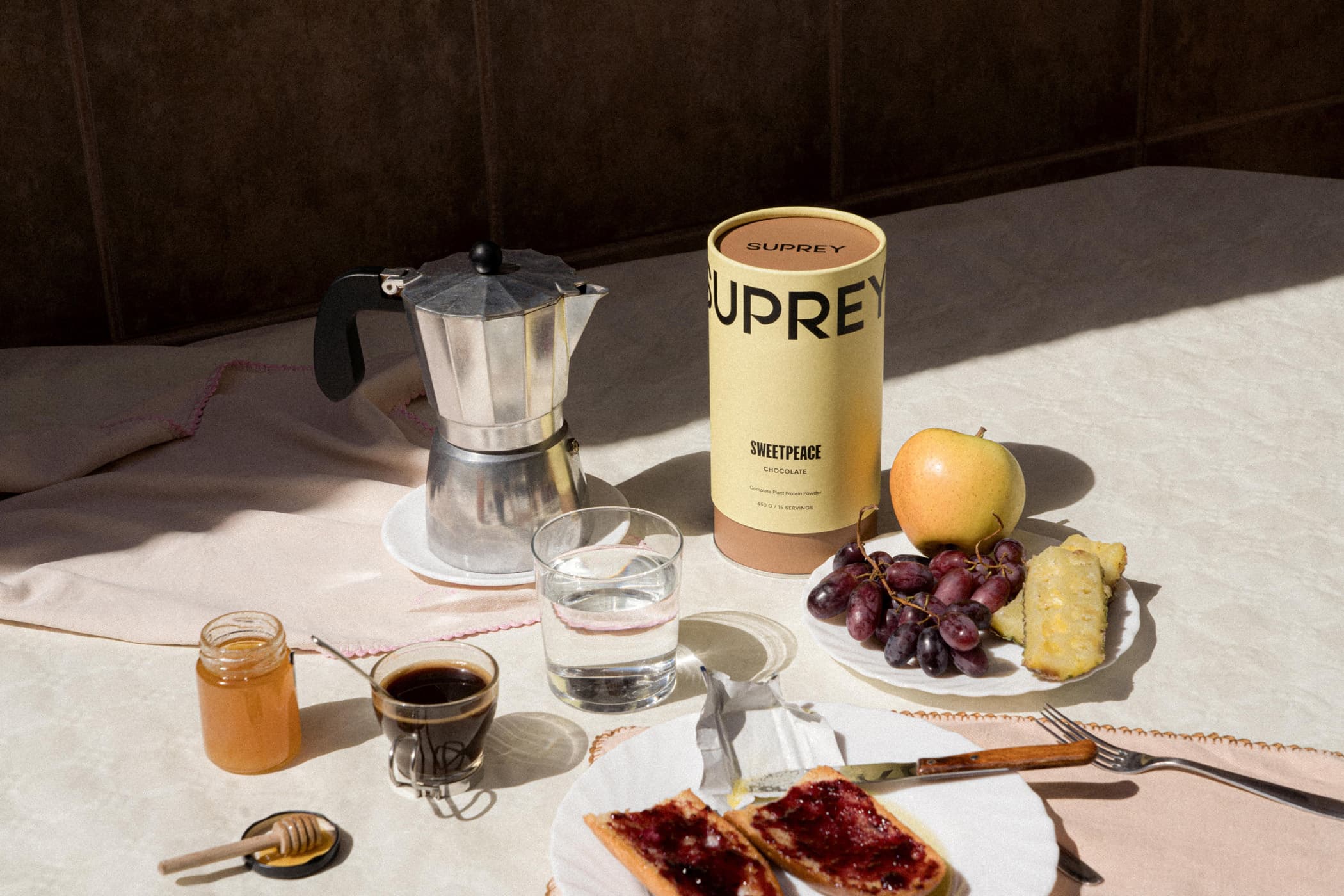 Breakfast still life with Suprey Sweetpeace Chocolate canister, moka pot, espresso, honey, fruit and jam toast in warm sunlight.